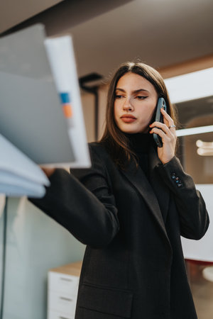 Professional woman in a black blazer talks on the phone while reviewing documents in a modern office