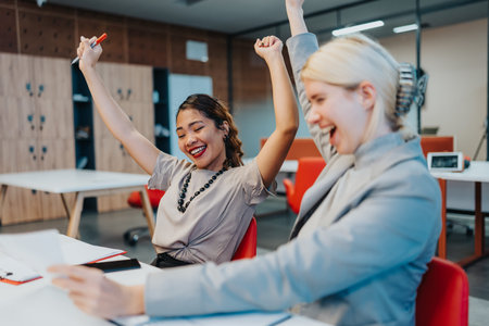 Two colleagues celebrate a win in the office, raising arms and smiling at their deskの写真素材