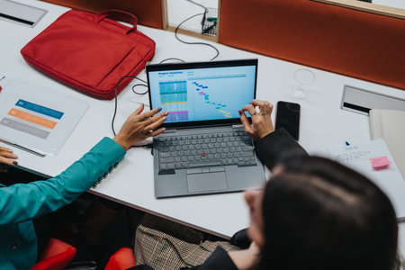 Business colleagues work on a laptop at a shared desk with notes and a red bag nearby
