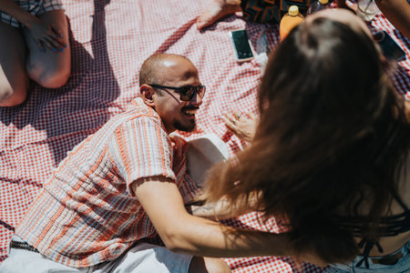 Relaxed man with sunglasses laughing at an outdoor picnic with friends on a checkered blanket