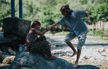 Friends enjoying a sunny summer by the river, helping each other balance on a rockの写真素材