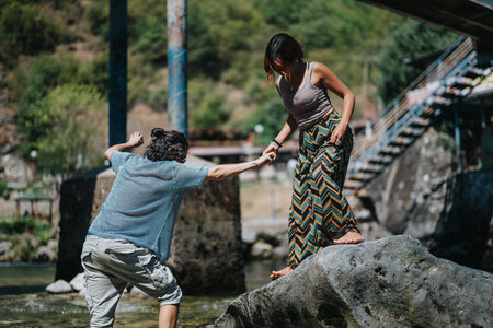 A man helps a woman balance on a rock by the river during a sunny outdoor momentの写真素材