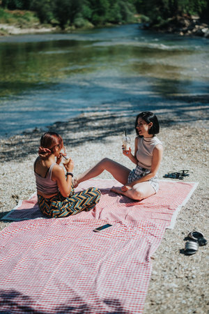 Two friends enjoy a sunny riverbank picnic on a red checkered blanket by the waterの写真素材