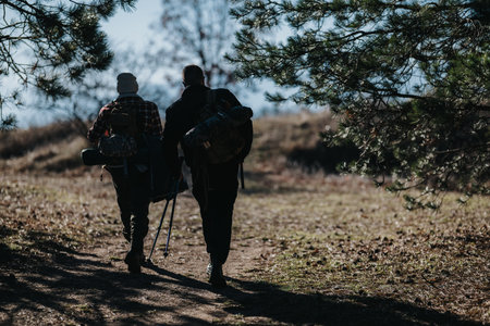Two hikers with backpacks walk along a forest trail under trees on a sunny day