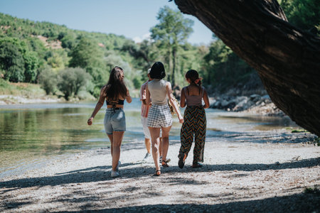 Four friends walk along a pebbled riverside on a sunny day, enjoying a casual summer outingの写真素材