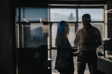 Two business professionals converse in a dim office with sunlit blinds and silhouettes