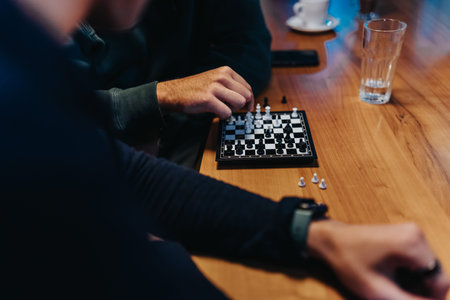 Two friends play a chess game on a wooden table with glass and cups nearby