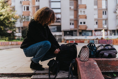 Crouching person with bags in an urban plaza, ready to travel, surrounded by backpacks and luggage