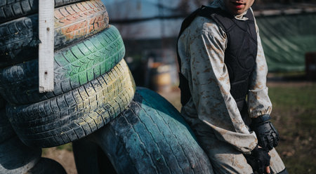 Paintball player in protective gear rests beside stacked tires during outdoor paintball game
