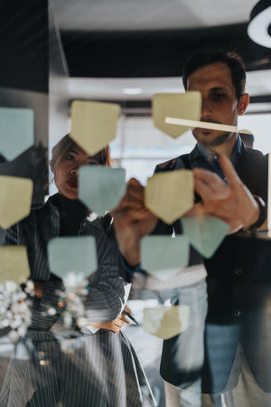 Two professionals review sticky notes on glass wall during a brainstorming session in a modern...