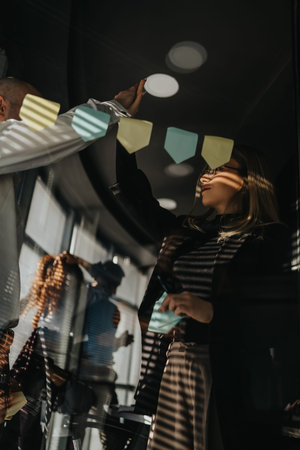 Team of professionals placing sticky notes on glass wall during a collaborative office projectの写真素材