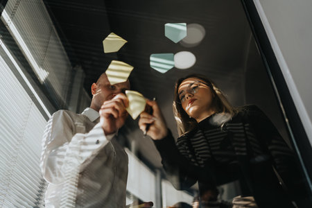 Two professionals brainstorm sticky notes on a glass wall during a business meetingの写真素材