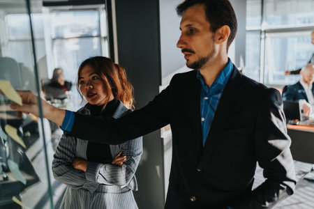 Professional man in suit presents notes on glass board while colleague observes in modern officeの写真素材