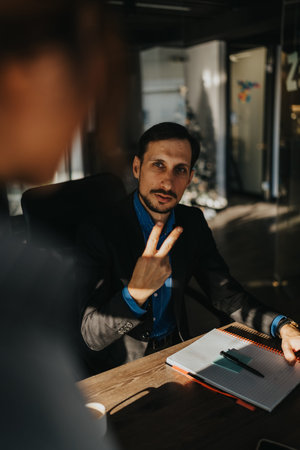 Professional man in a dark suit at a conference table with notebook and pen, making a peace sign...