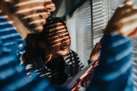 Two coworkers review documents near a sunlit window with striped blinds in an office setting