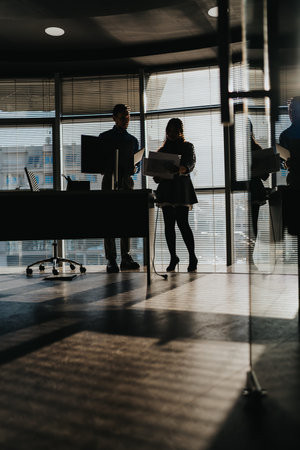 Two business people review documents in a modern, backlit office with blinds and glass wallsの写真素材