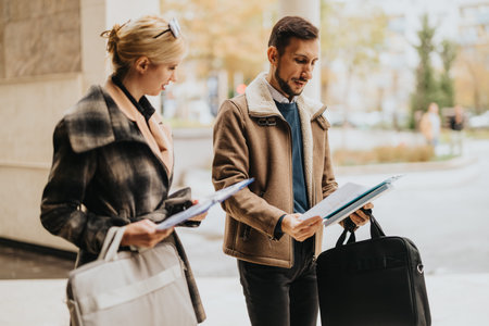 Two professionally dressed colleagues review documents outdoors with briefcases and folders