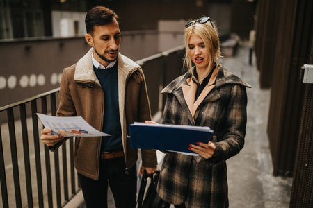 Two professionals discuss documents outdoors on a city walkway, reviewing files and charts together