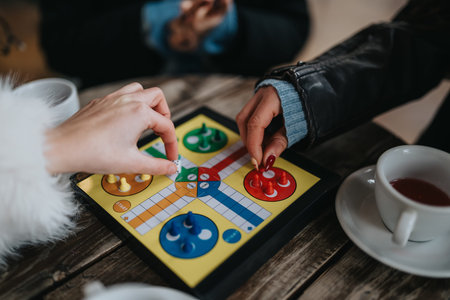 Girls play Ludo on a colorful board at a cafe table with coffee and jackets