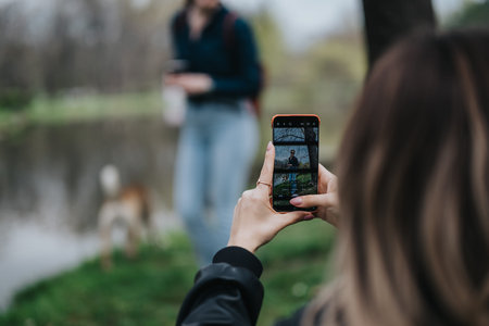 Woman takes photo of friend outdoors in park using smartphone, with a dog nearby watching