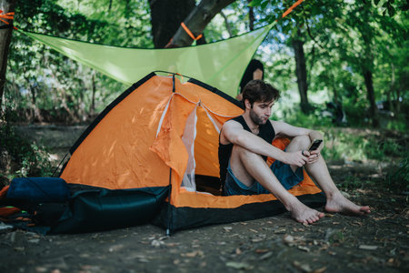 Man relaxing at a campsite outside an orange tent with smartphone in handの写真素材