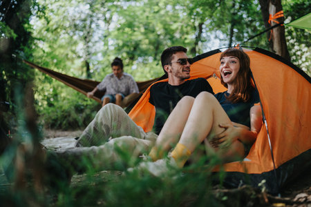 Happy couple relaxes in orange tent at forest campground with friends nearbyの写真素材