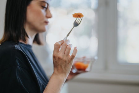 Young woman eating fresh fruit from a glass bowl with a fork by the window