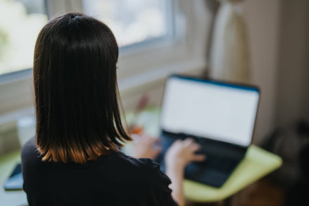 Woman working on a laptop at a home desk looking out the window while typing with focus