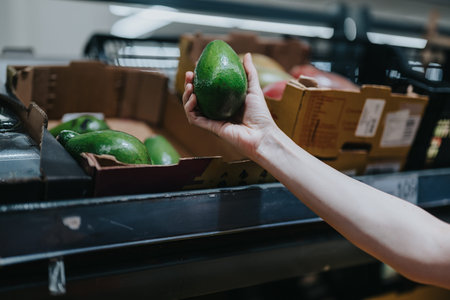 Woman picking a fresh avocado from a supermarket produce shelf while shopping for groceries