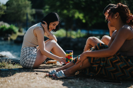 Three friends enjoy outdoor summer bonding by the river, playing a colorful block tower and laughing...