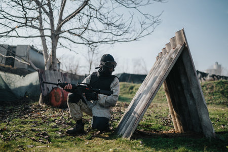 Tactical paintball player crouches behind improvised barrier with gun in outdoor field