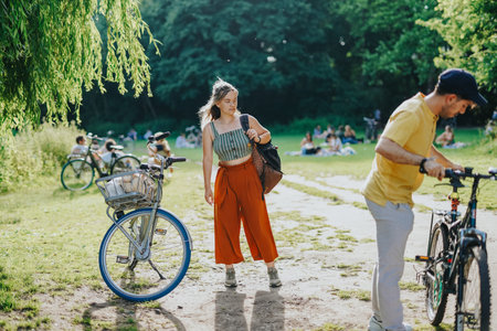 Young girl standing with bicycle in a sunny city park while people relax on the grass
