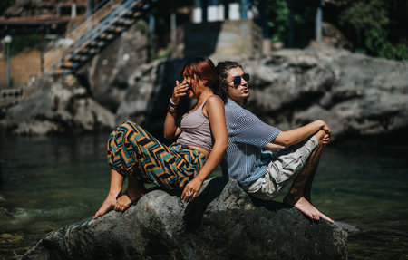 Two friends relax on a sunlit river rock, laughing together during a casual summer moment