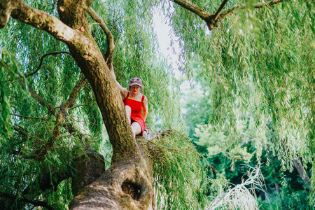 Girl in a red dress sitting on a weeping willow tree branch in a sunlit park
