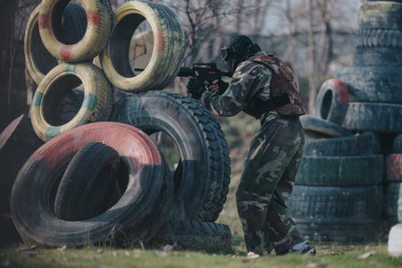 Camouflage paintball player hides behind stacked tires during outdoor game in a rugged field
