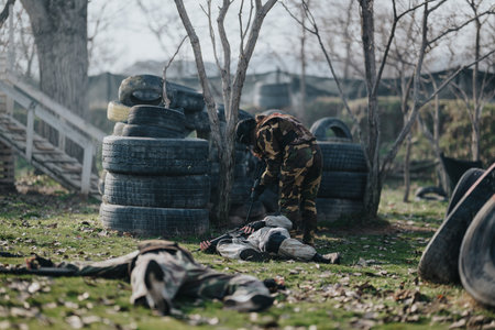Soldier in camo tending to injured teammate on paintball field with tires and trees outdoorsの写真素材