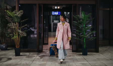 Young female tourist walking with suitcase through a glass entrance at nightの写真素材