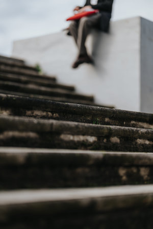 Person sitting on concrete ledge above weathered steps holding a red folder during an outdoor...