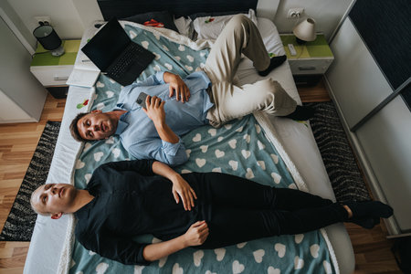 Two men relaxing on a bed at home with a laptop and smartphone in casual conversation