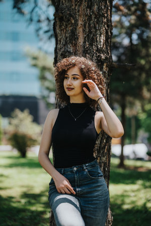 Woman with curly hair leaning against a tree in a sunny park, casual style and confident vibe