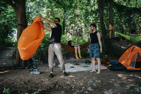 Friends setting up bright orange tent and hammocks in a forest campsite for outdoor recreation