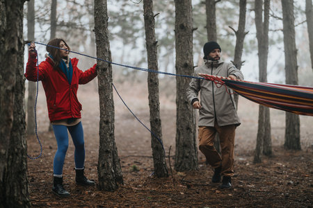 Two friends setting up a hammock in a misty pine forest during a cool outdoor camping trip