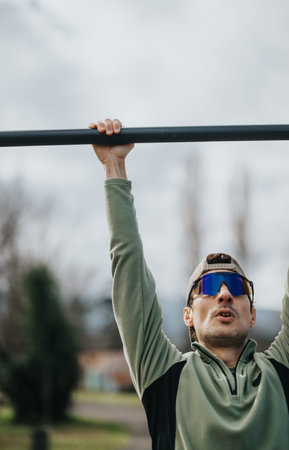 Man doing pull-up exercise outdoors wearing sunglasses and cap on a fitness bar