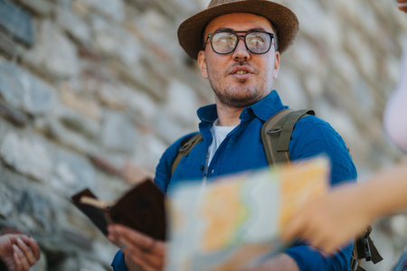 Male tourist with map and passport talking while wearing hat and backpack on stone street