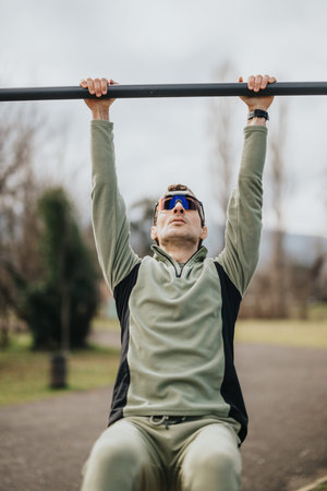Man doing an outdoor pull-up workout on a bar in a park wearing sunglasses and fleece sportswear