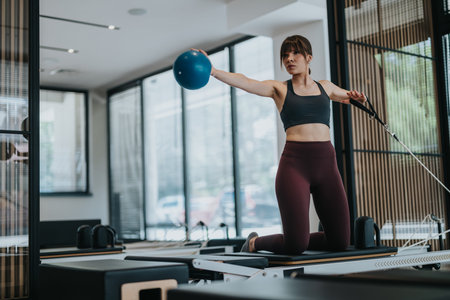 Young woman doing pilates reformer workout with medicine ball and resistance strap in studio