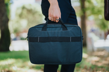 Person carrying a dark business briefcase by hand while walking outdoors in a park