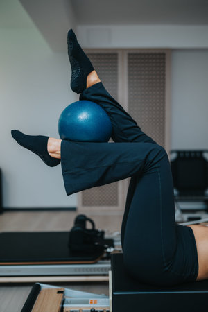 Pilates fitness workout on reformer with small exercise ball between crossed legs in studio