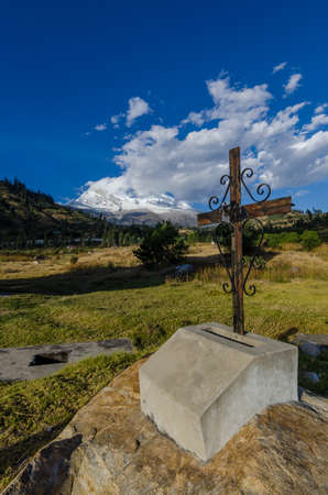 rusty cross on rock in yungay cemetery in foreground with snowy mountain in the background between cloudsの写真素材
