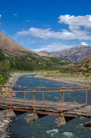 wooden suspension bridge held by ropes over river in valley surrounded by mountainsの写真素材
